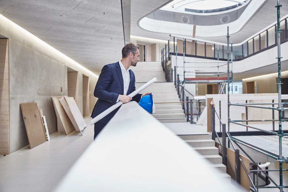 En välklädd arkitekt i mörk kostym och blå bygghjälm står vid ett vitt räcke i en modern kontorsbyggnad under konstruktion