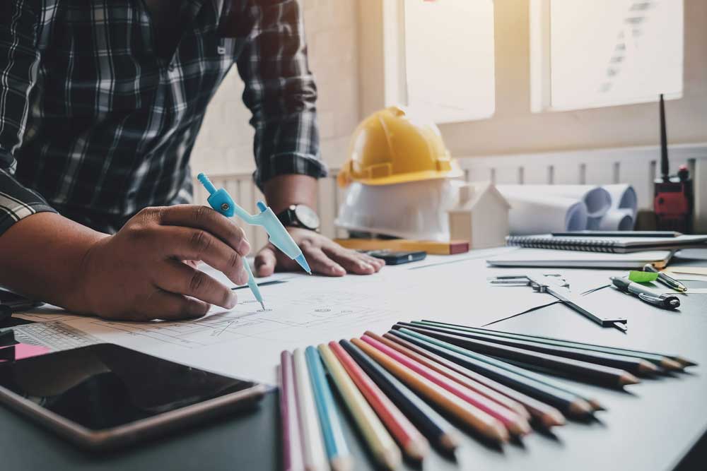 En arkitekt arbetar koncentrerat vid sitt skrivbord med en ritning framför sig, använder en ljusblå penna för att mäta och planera
