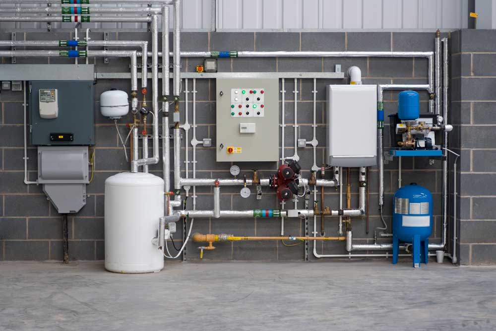 Serviceutrymme i en nybyggd fabrik visar ett komplett värmesystem med gaspanna, varmvattenberedare, kontrollpanel med varningsskyltar och symboler, samt elmätare