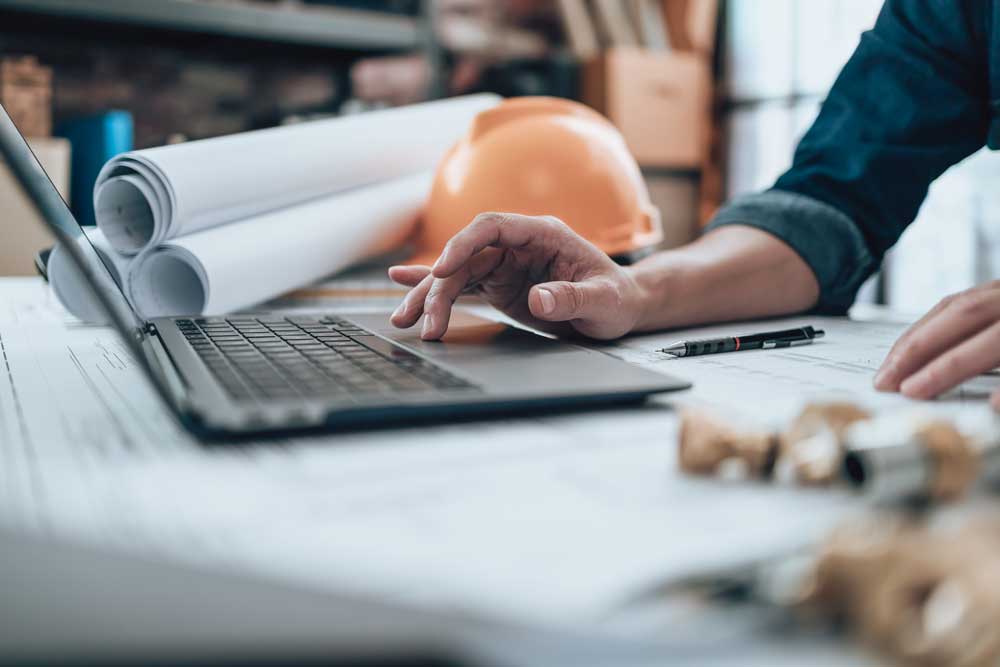 En ingenjör eller arkitekt arbetar vid en laptop omgiven av tekniska ritningar och verktyg på kontoret