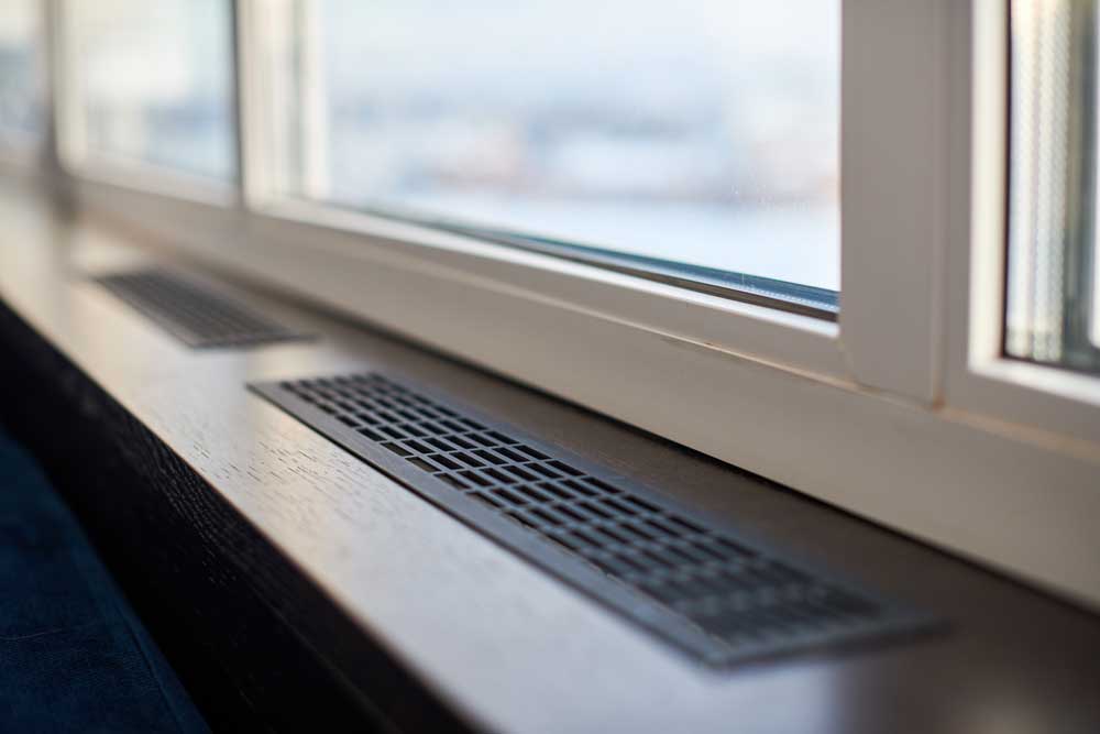 Ett modernt ventilationsgaller för värme installerat på en vit fönsterbräda framför ett tvåglasfönster